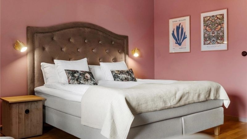 Elegant hotellrum p� Karlskrona Stadshotell med rosa v�ggar, stoppad s�nggavel, dubbels�ng med vita s�ngkl�der och dekorativa kuddar, samt stilrena tavlor p� v�ggen.